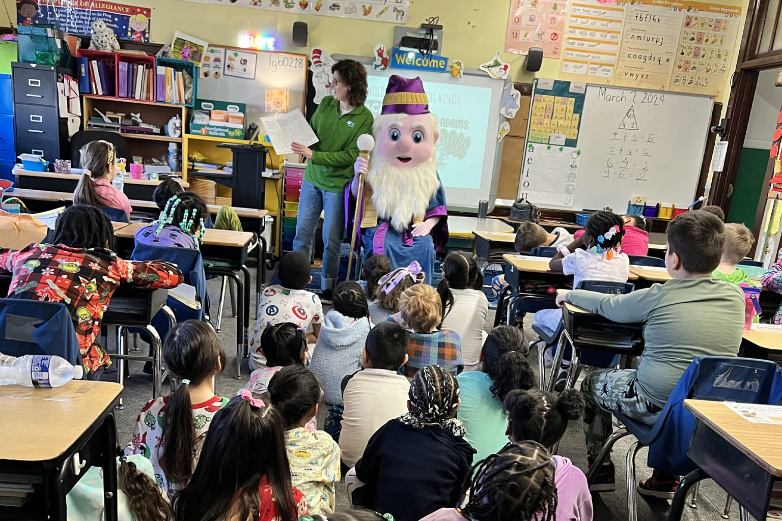A person dressed as a wizard reading to a group of first grade students.